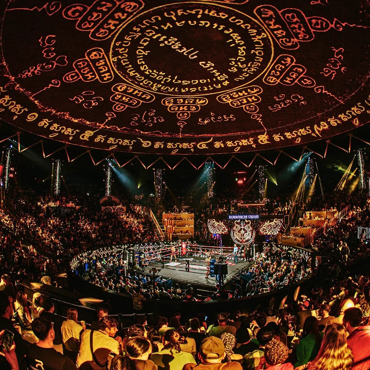 A packed Rajadamnern Stadium at night, with the ring lit up under a dome of glowing Sak Yant designs